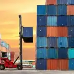 A red forklift moves containers in a busy shipping yard during sunset for logistics and trade.